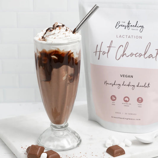 Lactation hot chocolate in a glass with whipped cream and a bag of The Breastfeeding Lactation Hot Chocolate mix on a white background.