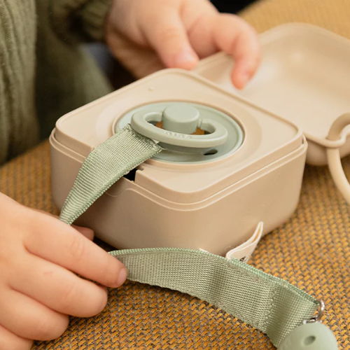 Close-up of a Ivory dummy box with sage dummy and dummy clip with strap, held by baby hands on a textured surface.