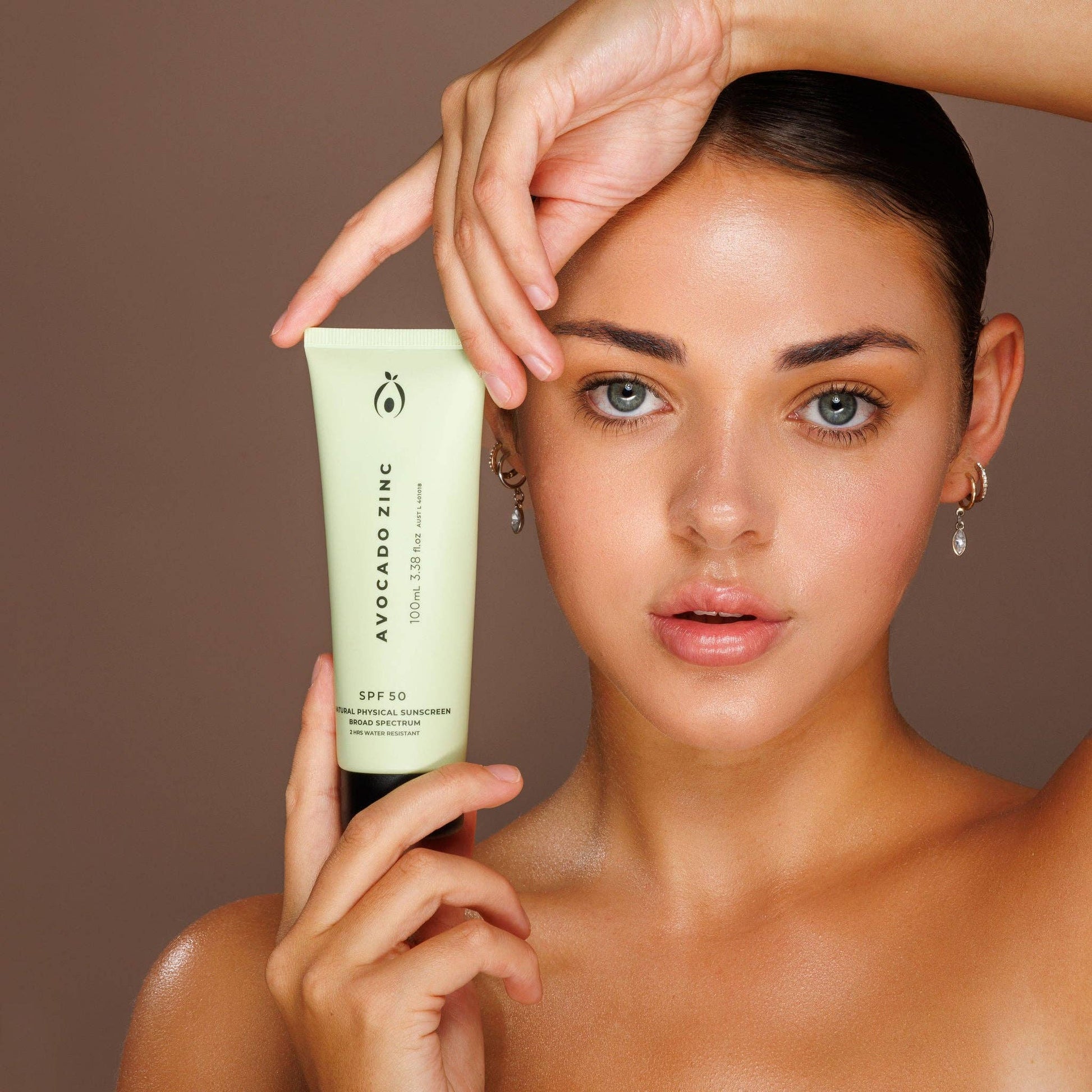 Woman holding a tube of Avocado Zinc sunscreen against a brown background