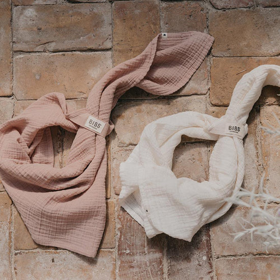 Two baby swaddles tied like bibs, one blush pink and one vanilla white, on a bricked surface.