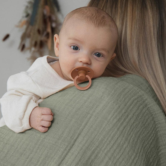 Lightweight sage BIBS muslin cloth for babies. Baby with dummy being cuddled by mum.