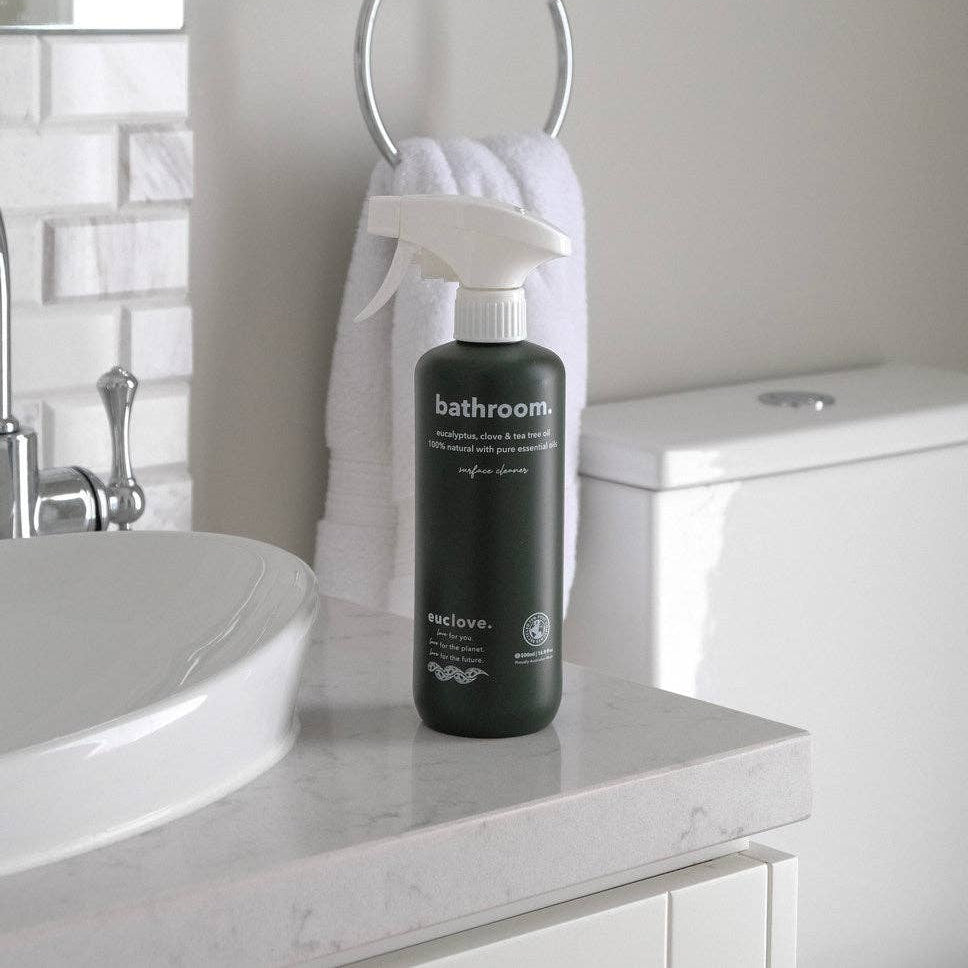 Black spray bottle labeled 'bathroom' on a bathroom counter with a sink and towel in the background.