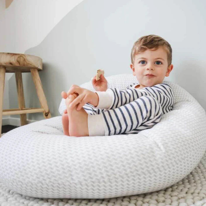 Child sitting on a textured body pillow chair in a room with a wooden stool and white wall.