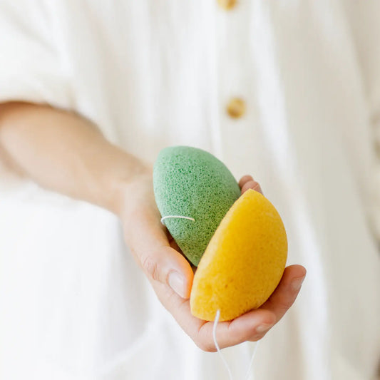 Hand holding a green and a yellow konjac sponge against a neutral background