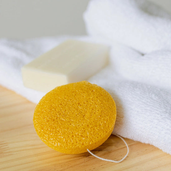 Yellow konjac sponge on a wooden board with a bar of soap and white towel in the background.