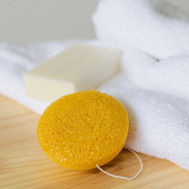 Yellow konjac sponge on a wooden board with a bar of soap and white towel in the background.