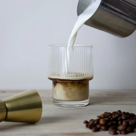 Pouring milk into the tumblr glass of coffee with coffee beans and a gold container on a wooden surface.