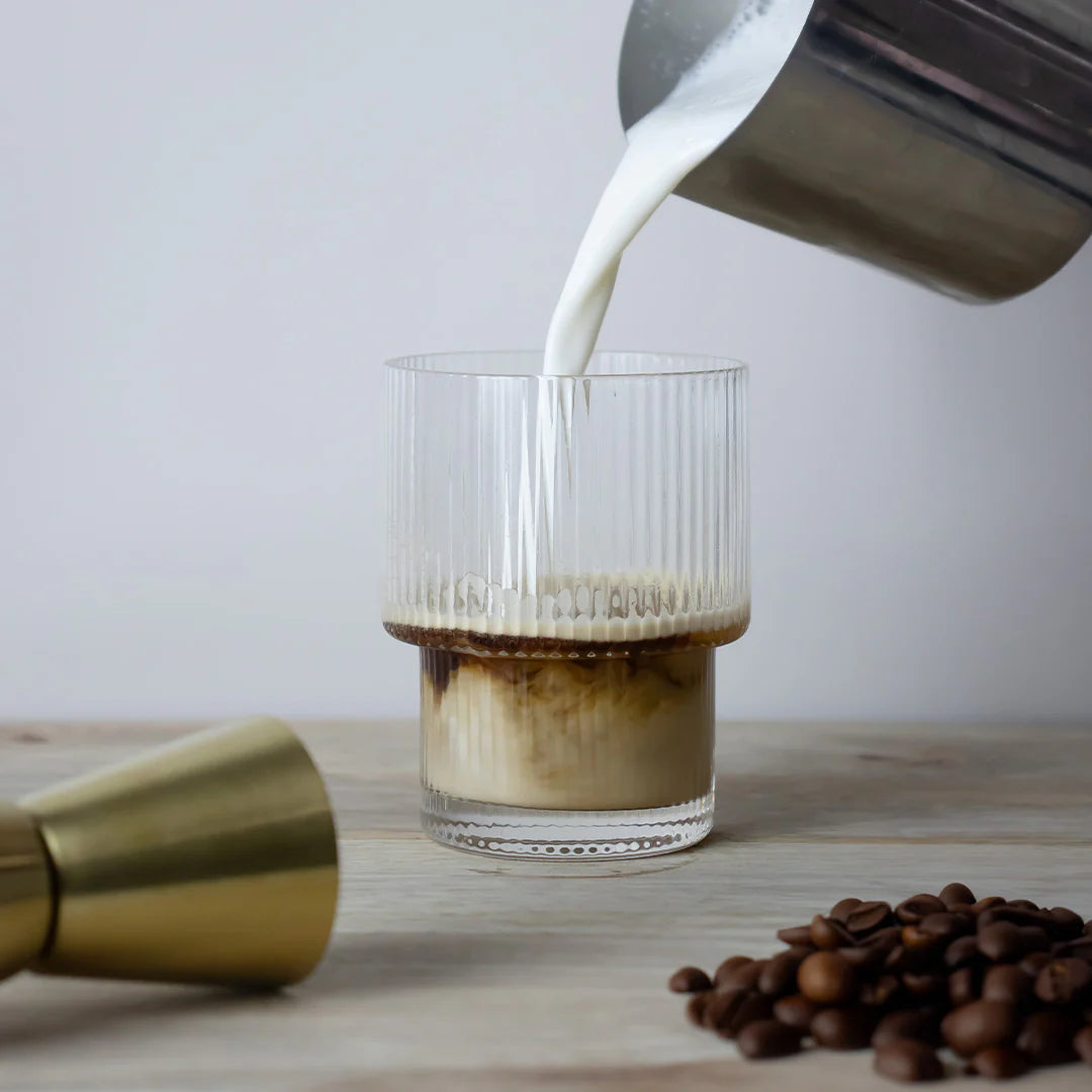 Pouring milk into the tumblr glass of coffee with coffee beans and a gold container on a wooden surface.