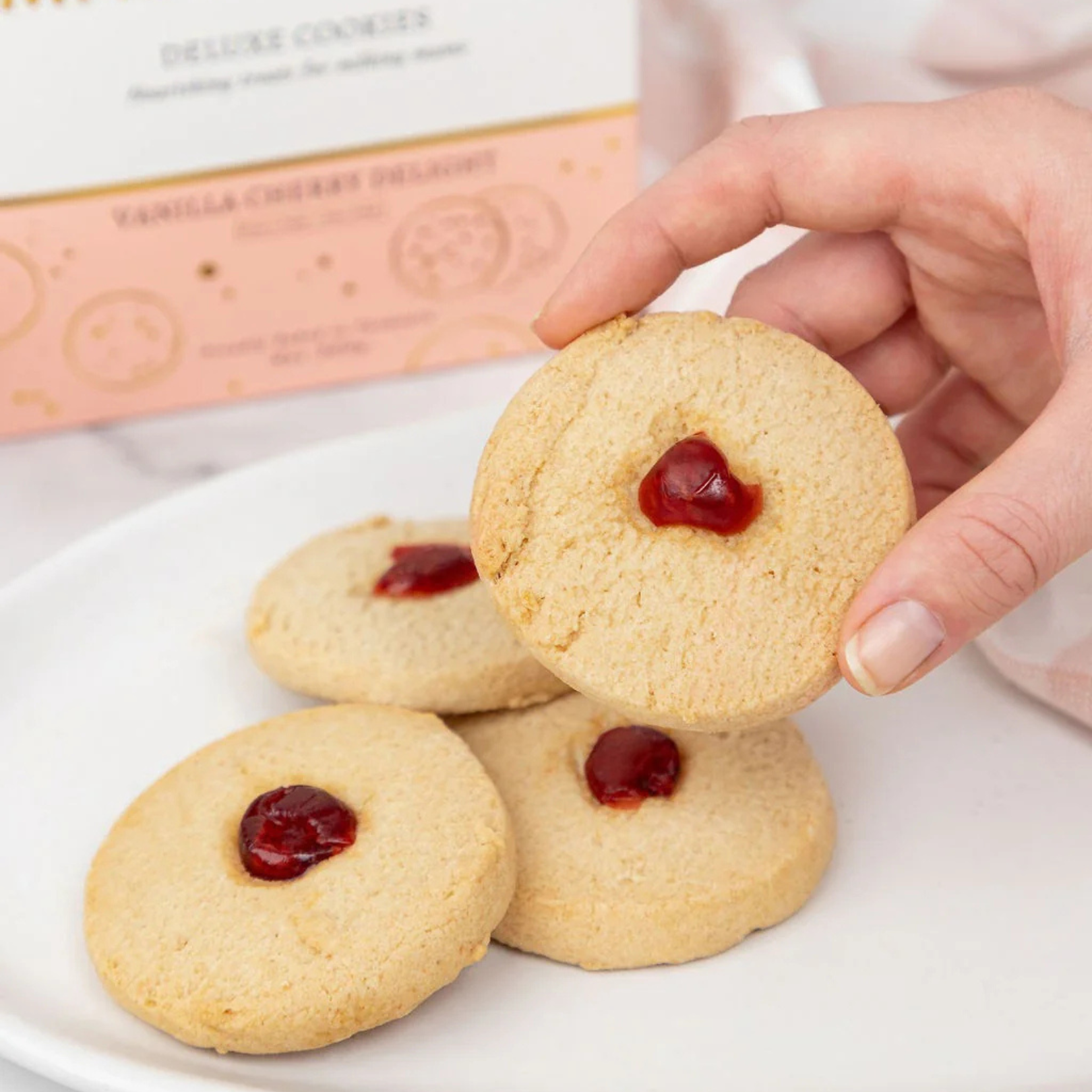 Hand picking a made to milk cookie with jam from a plate with a box in the background