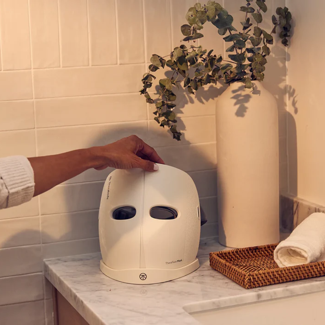White LED face mask on a marble countertop with a plant and vase in the background.