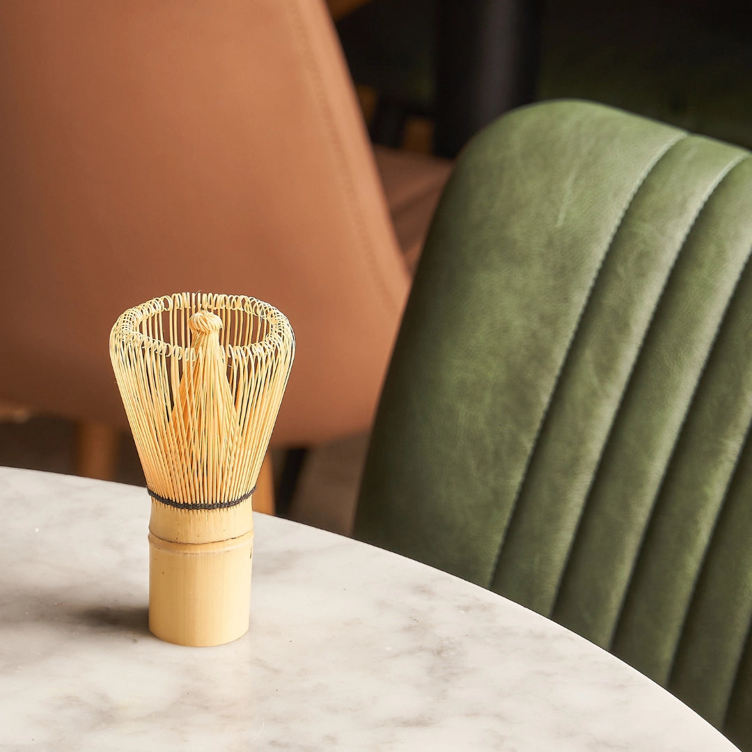 Bamboo matcha whisk on a marble surface with a blurred green chair and brown wall in the background