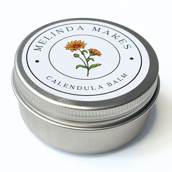 Metal tin with 'Melinda Makes' calendula balm with calendula floral on it on a white background
