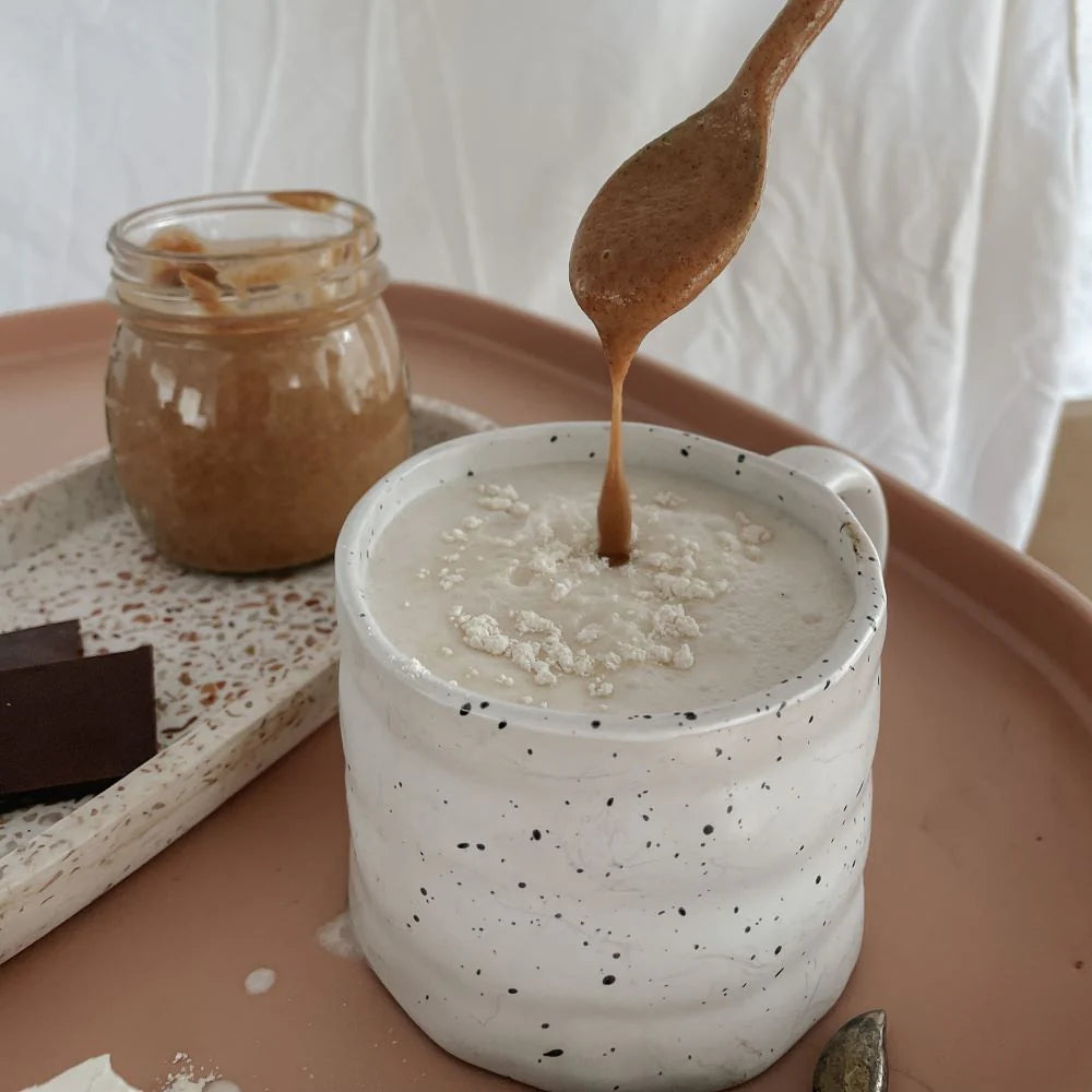 Spoon drizzling a caramel lactation liquid into a white speckled mug on a pink tray with a jar of the same liquid and a piece of chocolate.