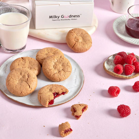 Lactation Raspberry Cookies with a bite taken out and raspberry filling, raspberries, and a glass of milk on a pink background with 'Milky Goodness' branding.  Dairy free and soy free