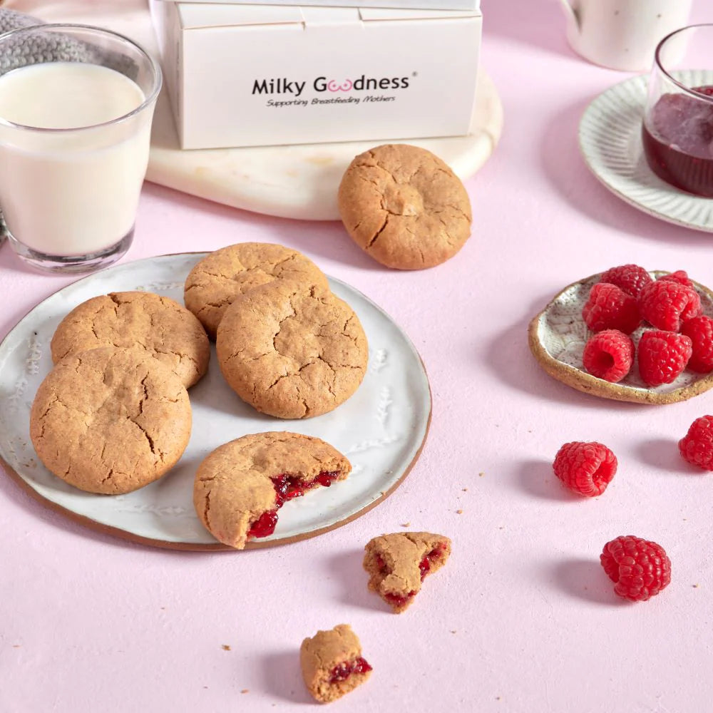 Lactation Raspberry Cookies with a bite taken out and raspberry filling, raspberries, and a glass of milk on a pink background with 'Milky Goodness' branding.  Dairy free and soy free