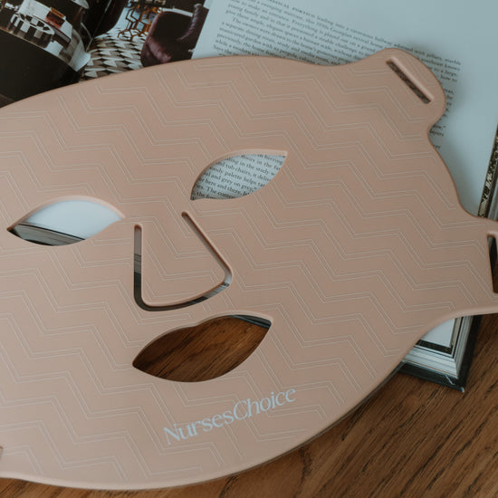 A pale pink silicone face mask template with a chevron pattern and the brand name 'NursesChoice' printed near the bottom, resting on a wooden surface over an open book.