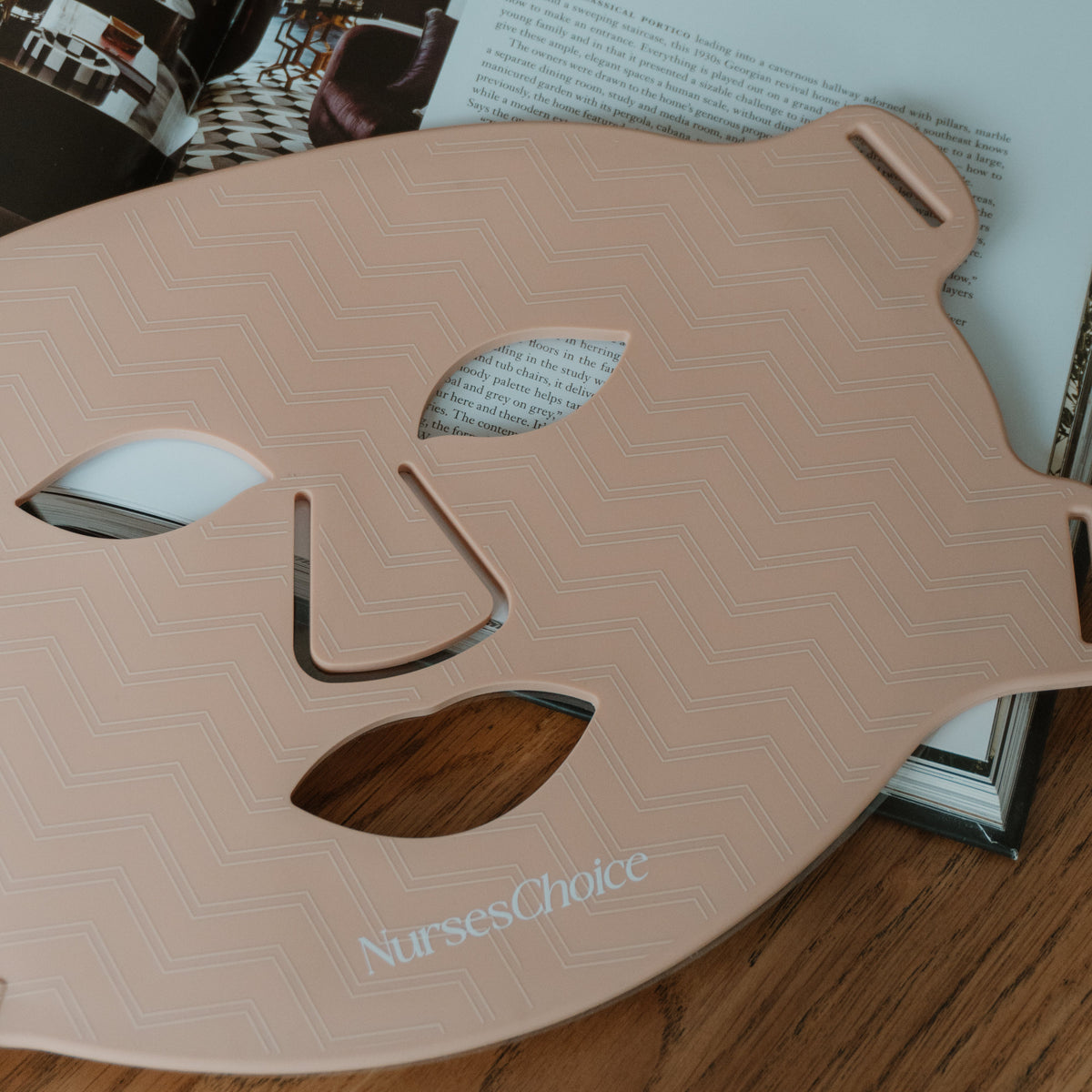 A pale pink silicone face mask template with a chevron pattern and the brand name 'NursesChoice' printed near the bottom, resting on a wooden surface over an open book.