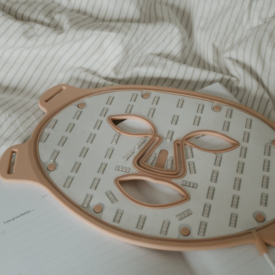 A rose gold/ nude and white sheet mask tray with a face-shaped cutout, resting on a white notebook over a beige and white striped duvet.