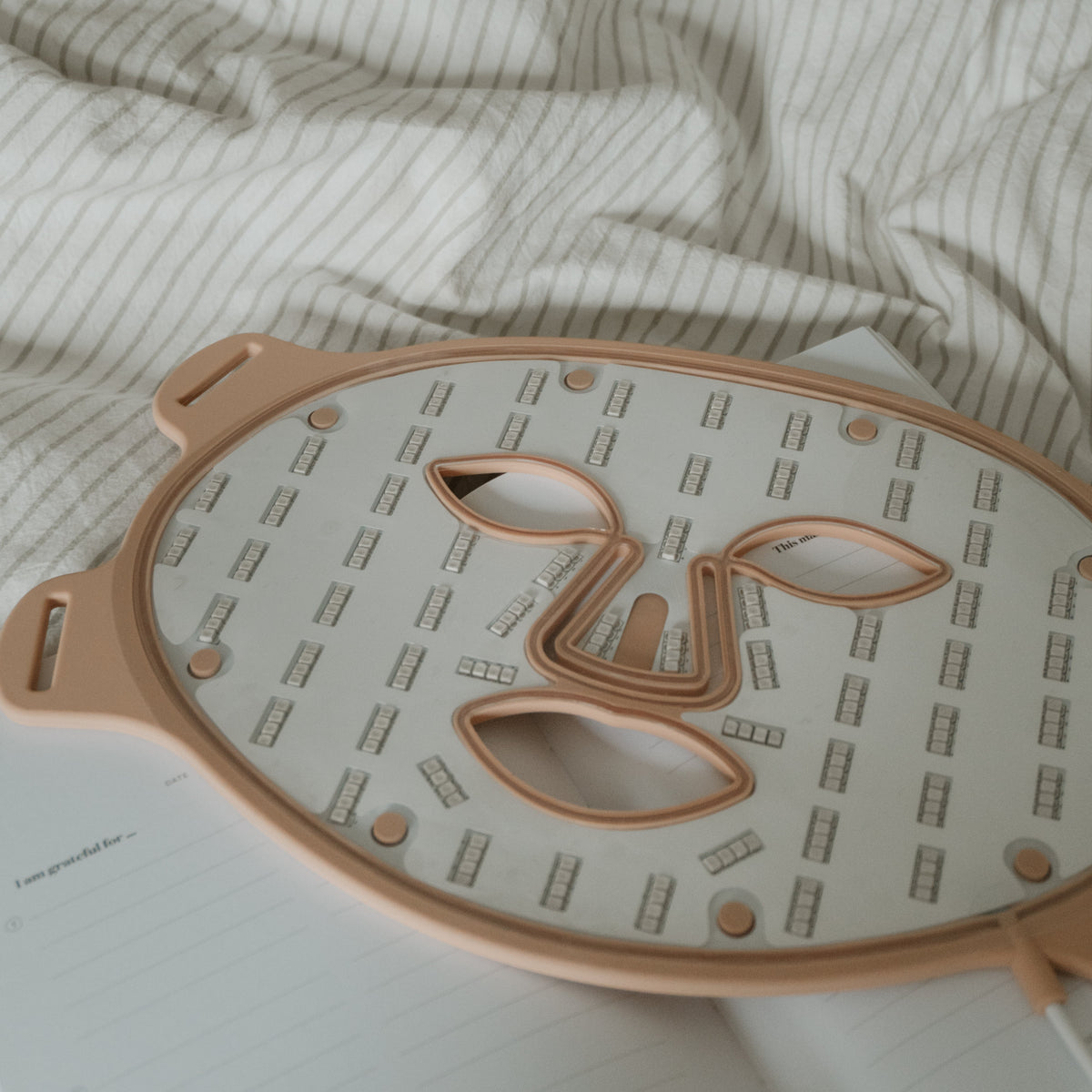 A rose gold/ nude and white sheet mask tray with a face-shaped cutout, resting on a white notebook over a beige and white striped duvet.