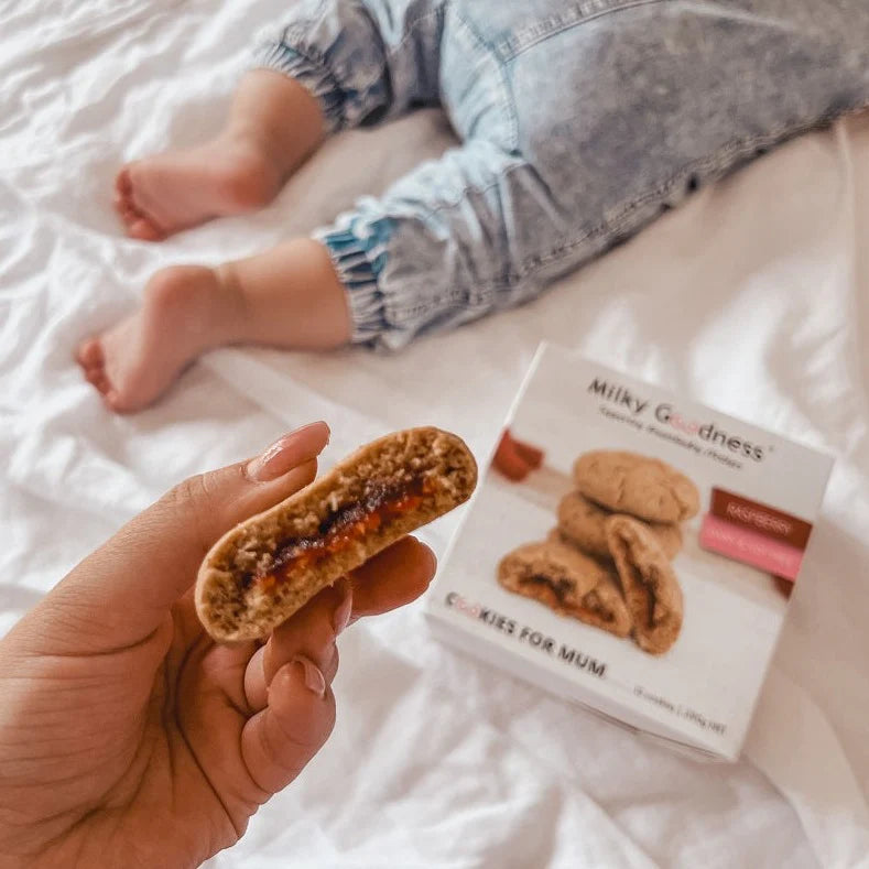 Person holding a half eaten lactation  dairy free and soy free raspberry cookie with 'Milky Goodness' packaging on a white surface with baby feet in background showing raspberry filling