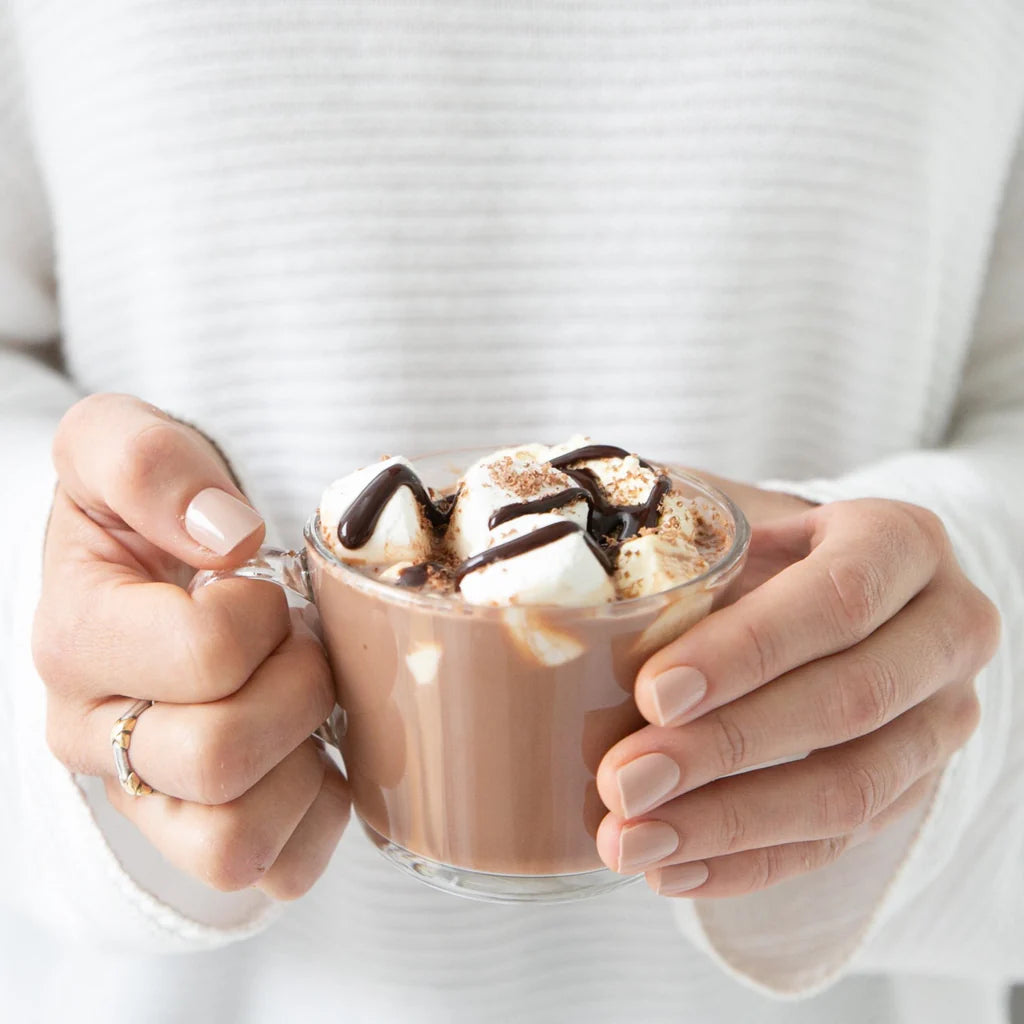 Person holding a glass mug of lactation hot chocolate with whipped cream and chocolate drizzle.