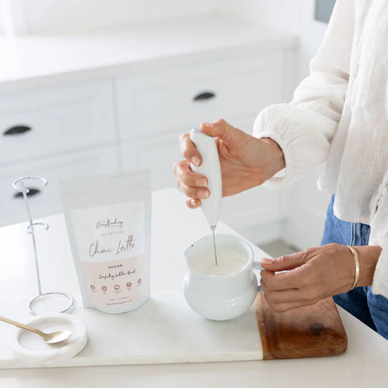 Person stirring a white cup with a spoon on a kitchen counter, next to a carton of 'Lactation Chai Latte' product. The breastfeeding co