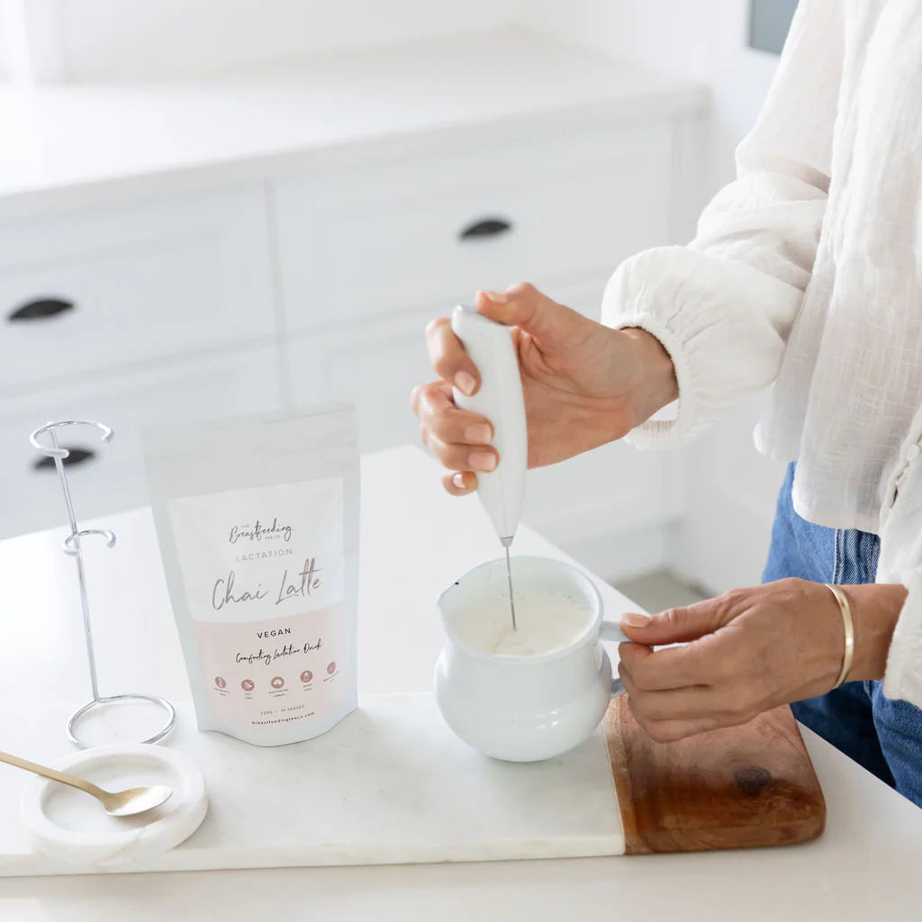 Person stirring a white cup with a spoon on a kitchen counter, next to a carton of 'Lactation Chai Latte' product. The breastfeeding co