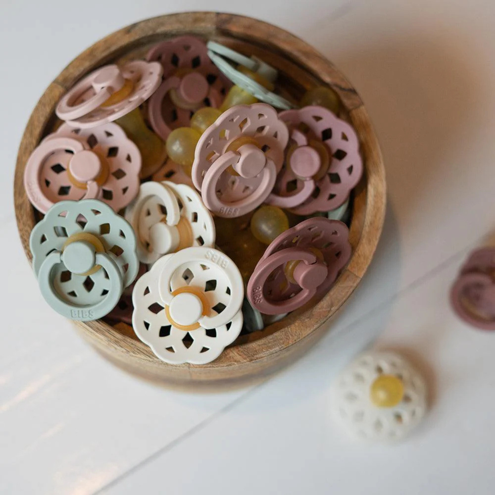 Wooden bowl filled with colorful bibs baby dummies on a white surface