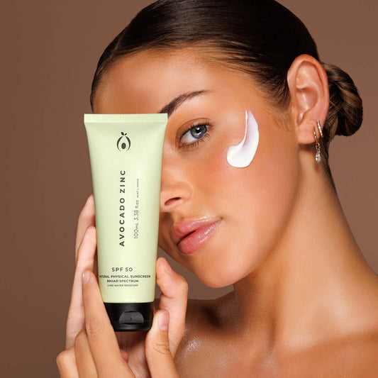 Woman applying sunscreen with a tube of Avocado Zinc on a brown background