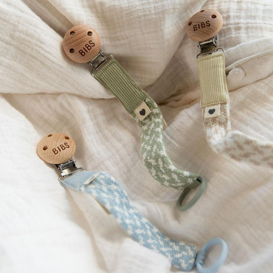 3 of dummy clips or pacifier clips. Light timber badge that says bibs on top of metal clip attached. Braided cotton bands in ivory and baby blue, ivory and sage and ivory and vanilla with dummy ring on ends.