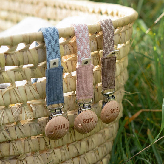 3 of dummy clips or pacifier clips. Hanging of woven basket. Light timber badge that says bibs on top of metal clip attached. Braided cotton bands in Cornflower and dusty blue, ivory and blush and dark oak and vanilla.
