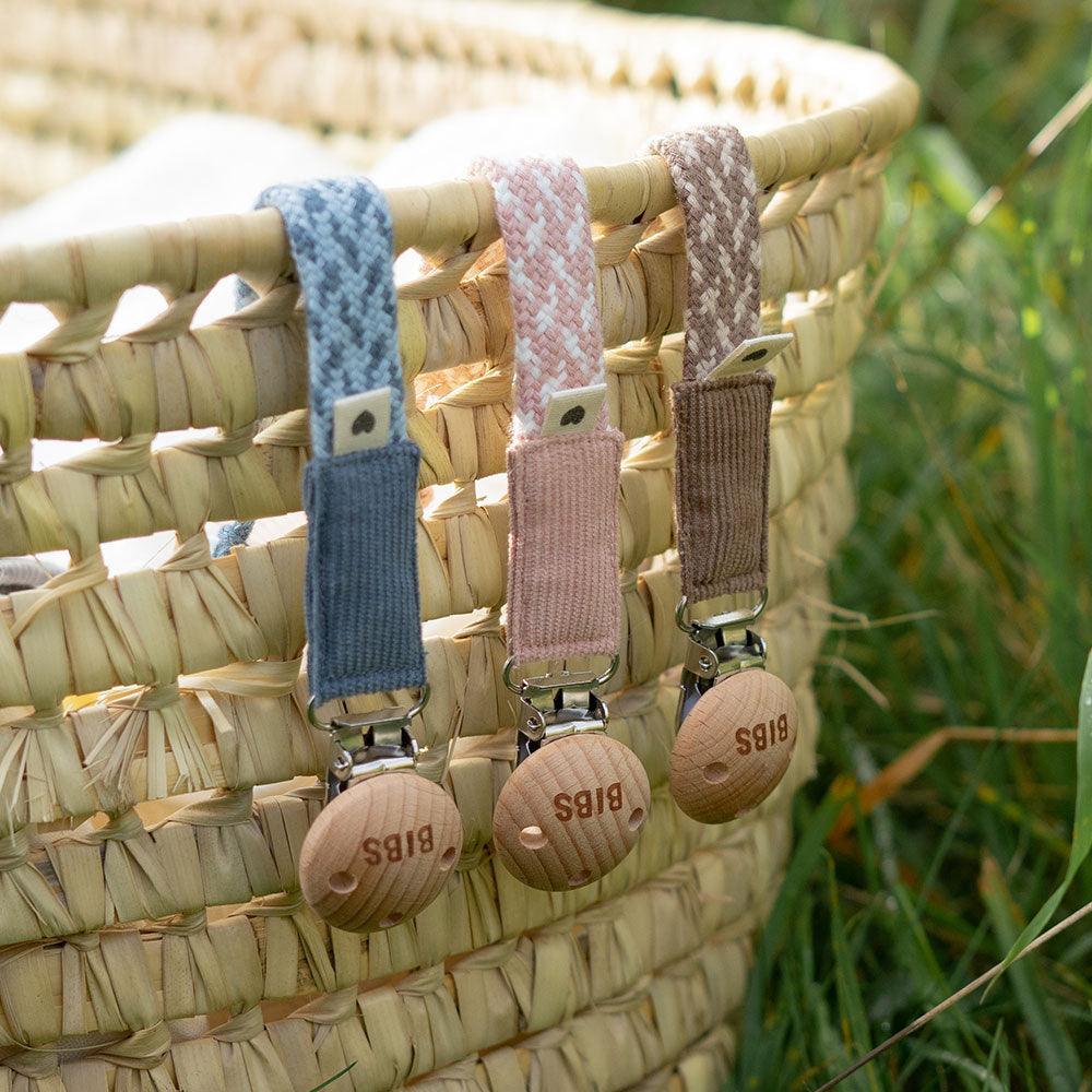 3 of dummy clips or pacifier clips. Hanging of woven basket. Light timber badge that says bibs on top of metal clip attached. Braided cotton bands in Cornflower and dusty blue, ivory and blush and dark oak and vanilla.