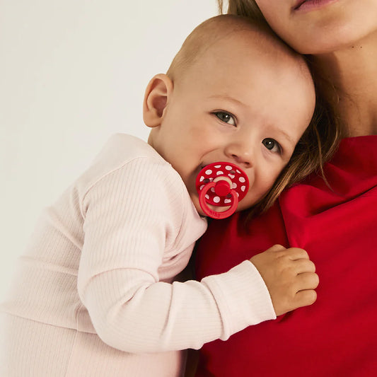 Baby with a red polka dot pacifier or dummy held by a person against a white background