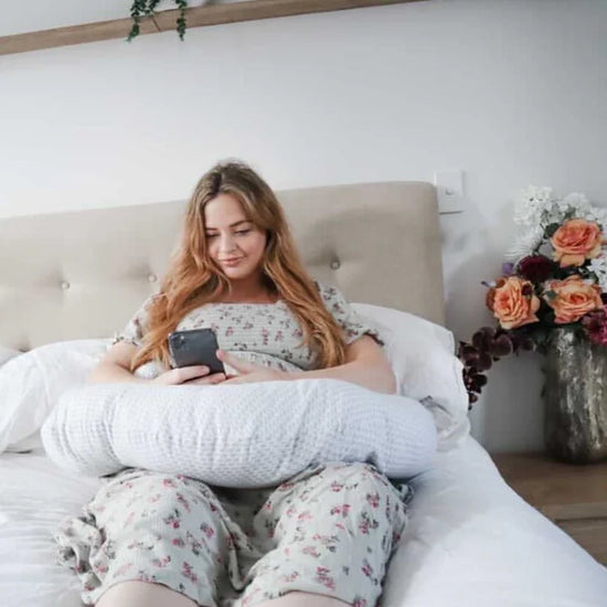 Woman sitting on a bed using a phone with a body pillow behind her back