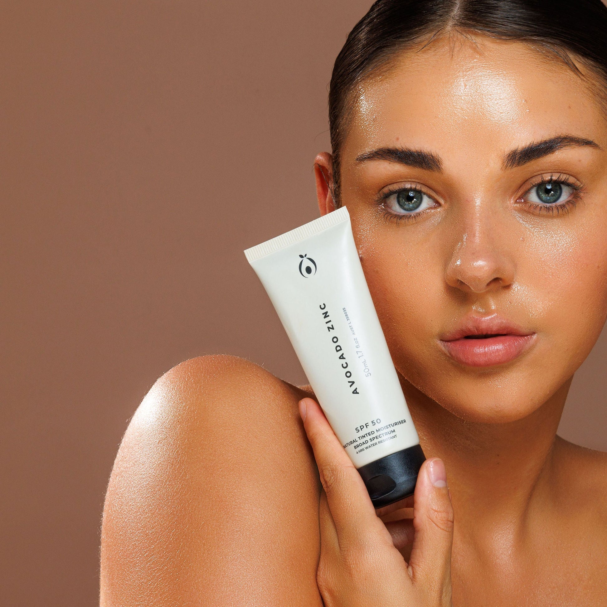 Woman holding a tube of Avocado Zinc sunscreen against a brown background