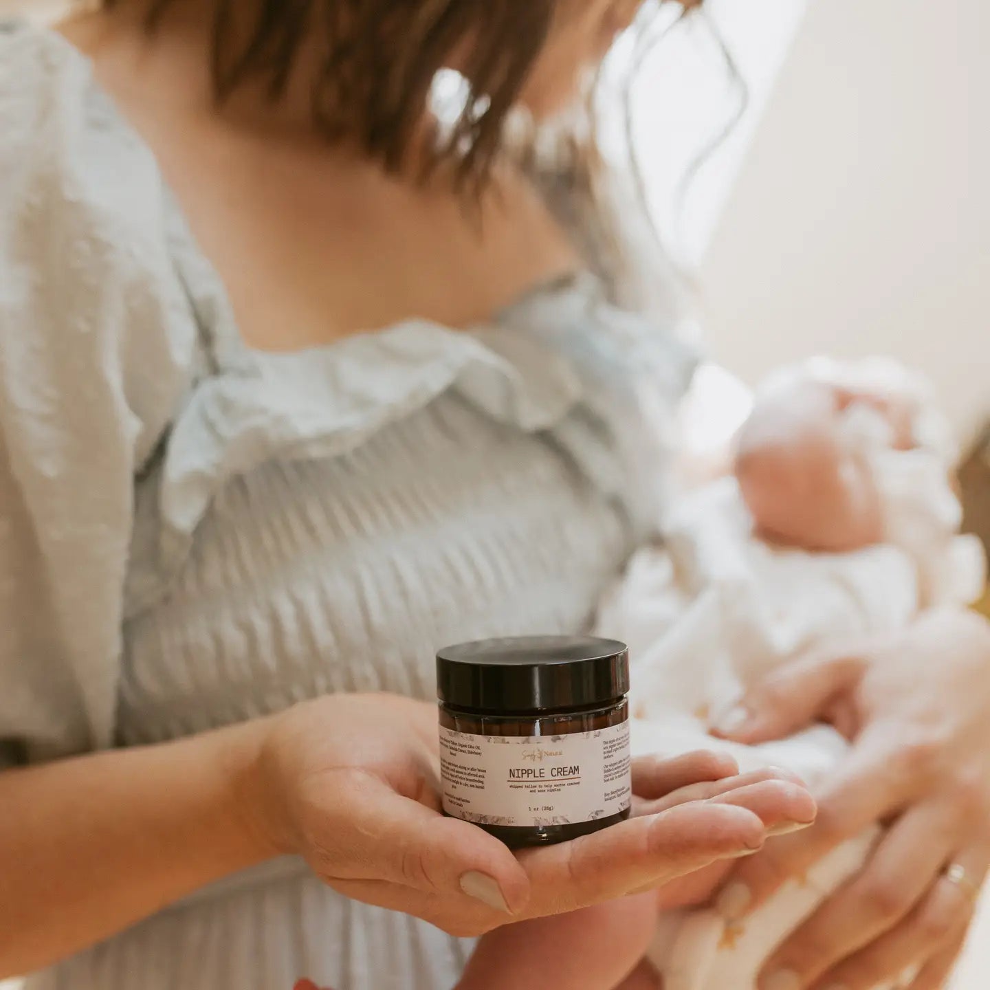 Person holding a jar of  nipple cream with a baby in the background