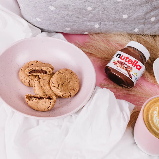 Nutella lactation Cookies on a pink plate with a jar of Nutella and a cup of coffee on a white surface.