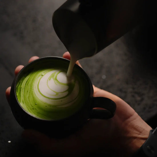 Person pouring a green matcha drink into a black mug with a decorative pattern.