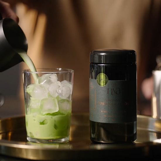 Green drink being poured into a glass with ice next to a bottle labeled 'Nyu' matcha on a reflective surface.