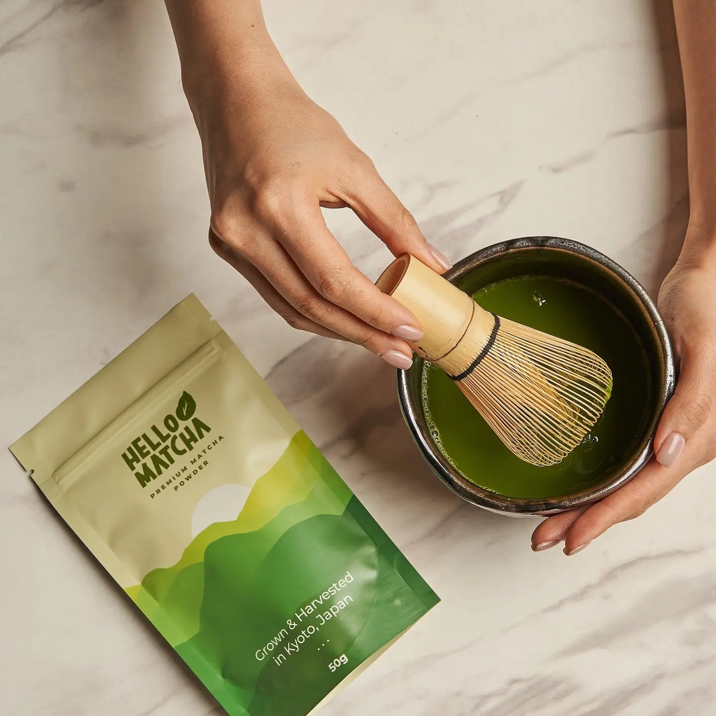 Person preparing matcha tea with a whisk and Hello Matcha packaging on a light surface.