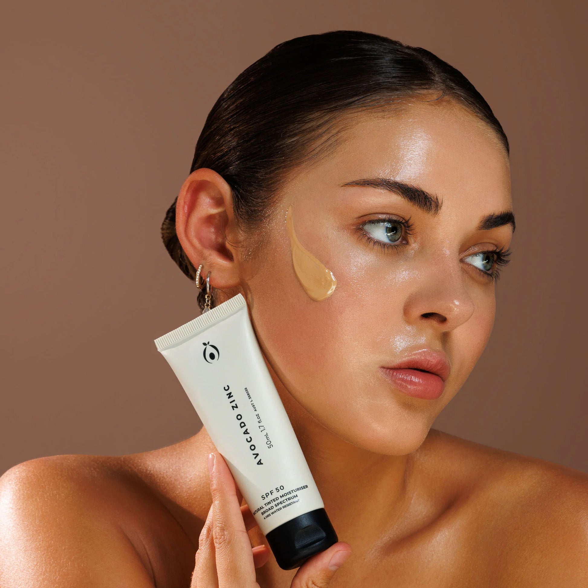 Woman applyed tinted moisturiser skincare product with a tube of Avocado Zinc on a beige background