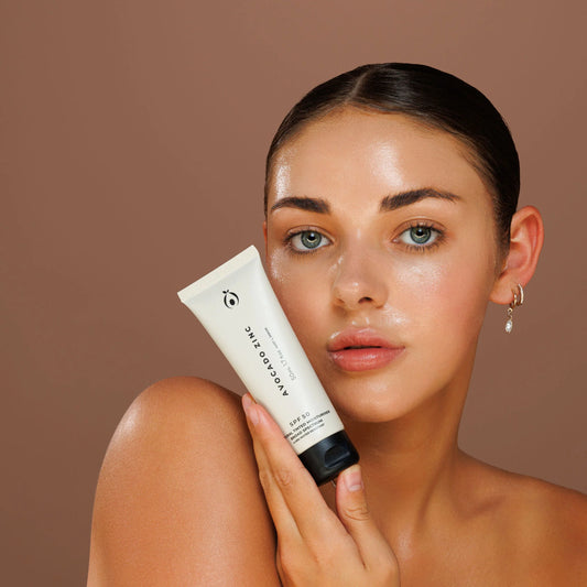Woman holding a avocado zinc tinted moisturiser skincare product against a brown background