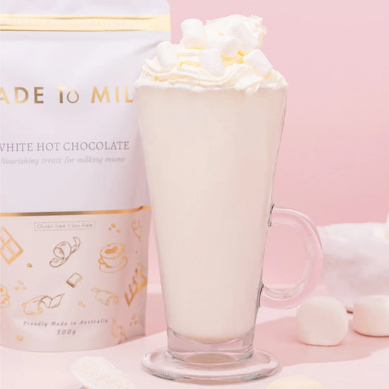 Glass of lactation white hot chocolate with whipped cream next to a branded container of made to milk on a pink background